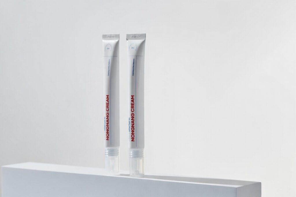 깨끗한 화이트 큐브 위에 두 개의 농낭크림 튜브가 나란히 세워져 있는 미니멀한 구성의 사진입니다. 화이트 톤의 깔끔한 배경이 제품의 청결함과 세련된 패키지 디자인을 돋보이게 합니다.